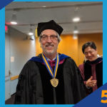 A photo of Duncan RyanMann at a graduation ceremony wearing black regalia and a medal.