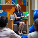 Photo of President Lisa Vollendorf smiling as she gestures to a book with a colorful mural in the background and individuals sitting in front of her