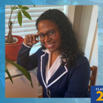 Robyn Guzman seated indoors at a desk near a potted plant, wearing a navy blazer and white shirt. A blue graphic border includes a torch logo at top left and the text “Class of 2026” at bottom right.