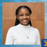 Formal portrait of Gabby Damis wearing a white long‑sleeve top and a heart pendant necklace, set against a wood-paneled background. A blue graphic frame surrounds the photo with a torch logo in the top left and the text “Class of 2026” in the bottom right.