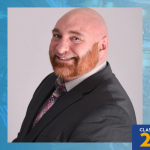 Formal studio portrait of Kevin DiPalma in a dark suit and tie. Blue background includes a shield-and-torch logo and text reading “Class of 2026.